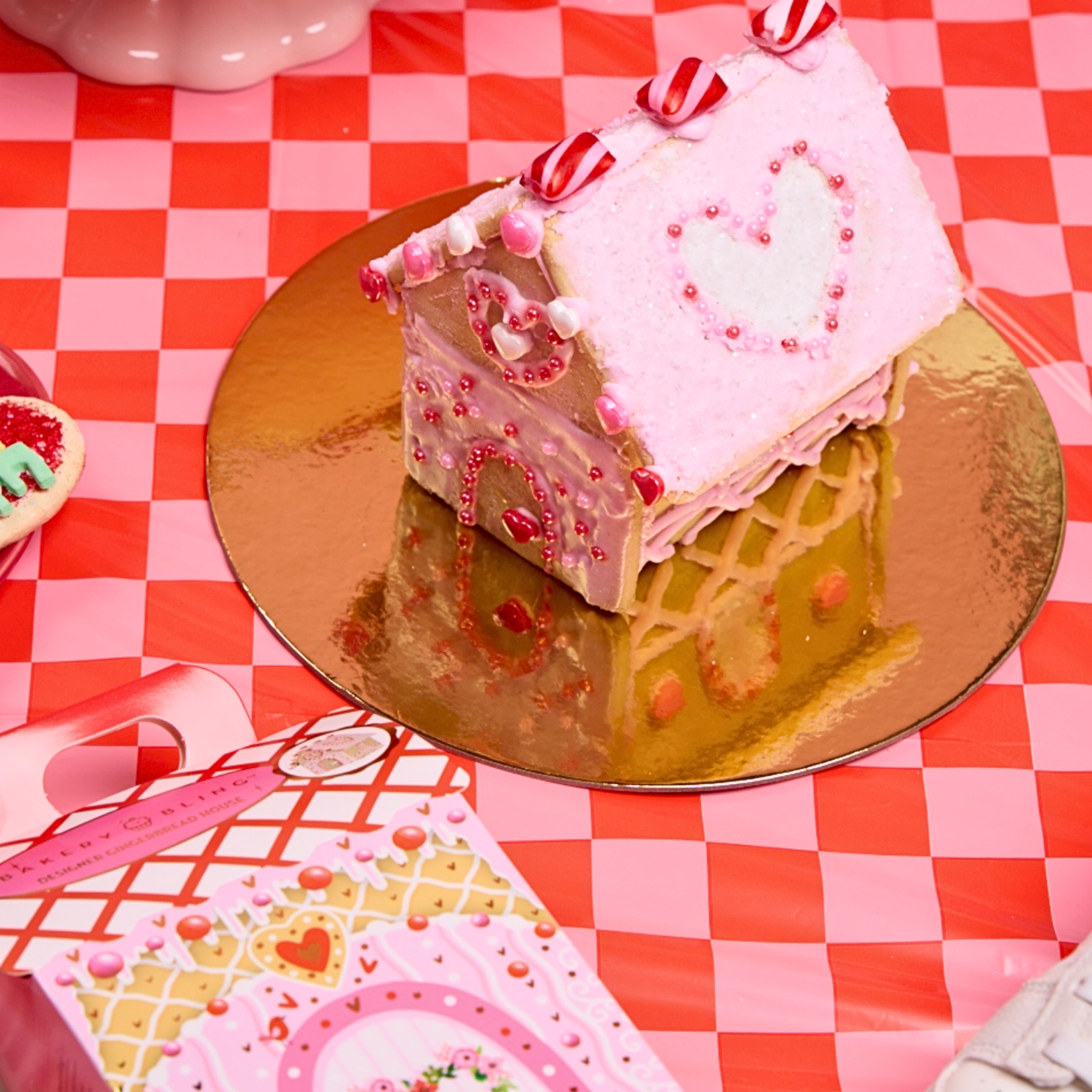 Mini Pink Valentine’s Day-themed cookie (gingerbread) house decorated with pink royal icing, red heart candies, and edible glitter, set on a gold platter with festive pink and red Valentine’s decor in the background